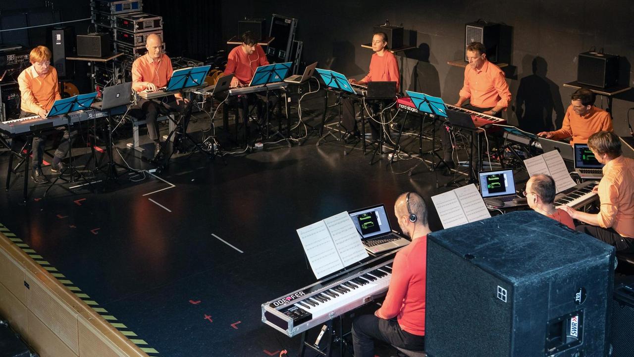 Neun Synthesizer-Spieler sitzen mit ihren Instrumenten in einem großen U, damit sie sich beim gemeinsamen Spiel sehen können. Neun Synthesizer-Spieler sitzen mit ihren Instrumenten in einem großen U, damit sie sich beim gemeinsamen Spiel sehen können.