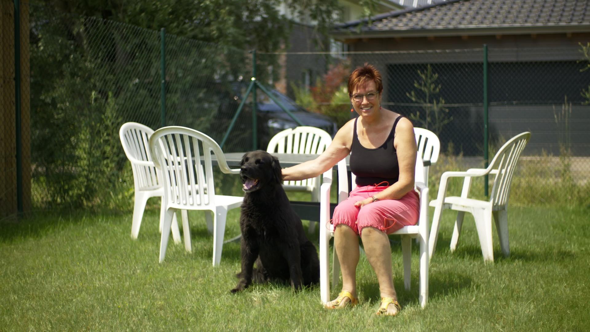 Szene aus dem Dokumentarfilm "Monobloc": Eine Frau sitzt im Garten auf einem weißen Plastikstuhl, um sie herum stehen weitere dieser Stühle. Die Frau streichelt einen Hund, der neben ihr auf dem Rasen sitzt. Szene aus dem Dokumentarfilm "Monobloc": Eine Frau sitzt im Garten auf einem weißen Plastikstuhl, um sie herum stehen weitere dieser Stühle. Die Frau streichelt einen Hund, der neben ihr auf dem Rasen sitzt.