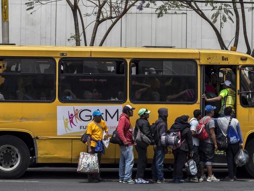 Mehrere Menschen mit Taschen und Rucksäcken stehen in einer Schlange am Eingang eines gelben Busses, der am Straßenrand vor einem Gebäude steht.