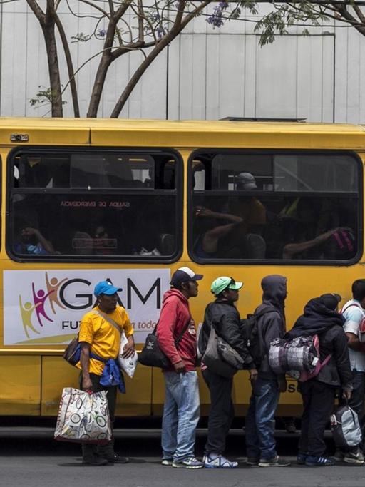 Mehrere Menschen mit Taschen und Rucksäcken stehen in einer Schlange am Eingang eines gelben Busses, der am Straßenrand vor einem Gebäude steht.