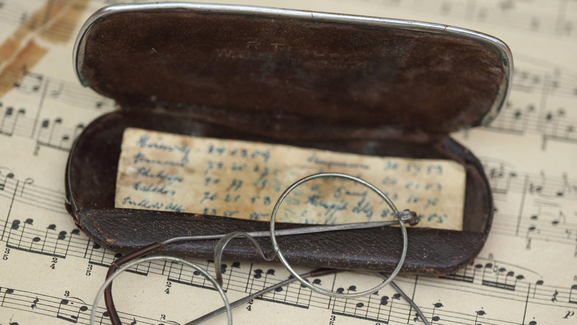 Eine Brille liegt mit einem Etui und handschriftlichen Notizen auf einem Notenblatt. Eine Brille liegt mit einem Etui und handschriftlichen Notizen auf einem Notenblatt.