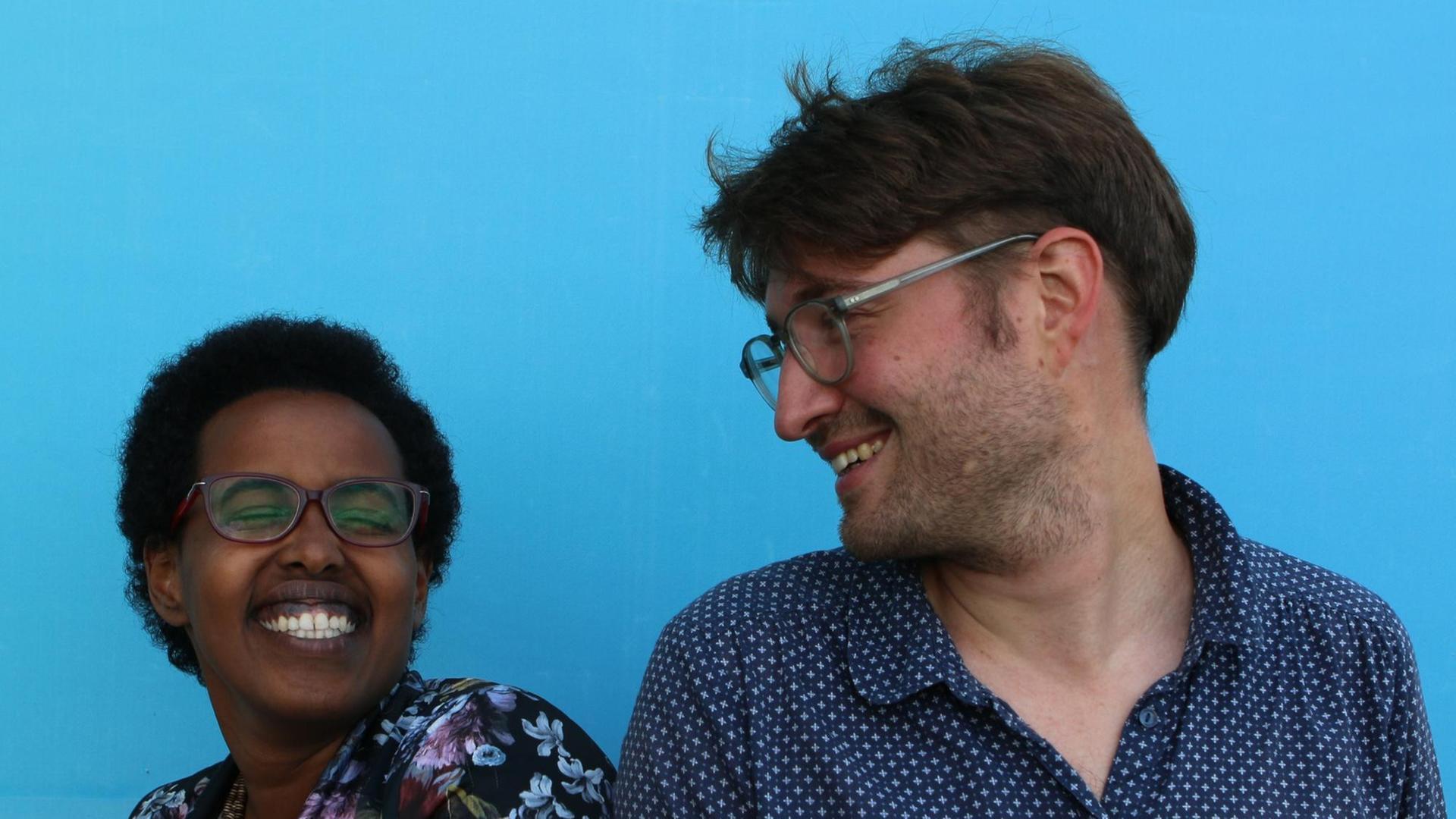 Sarah Fartuun Heinze und Kolja Unger stehen lächelnd vor einer blauen Wand Sarah Fartuun Heinze und Kolja Unger stehen lächelnd vor einer blauen Wand