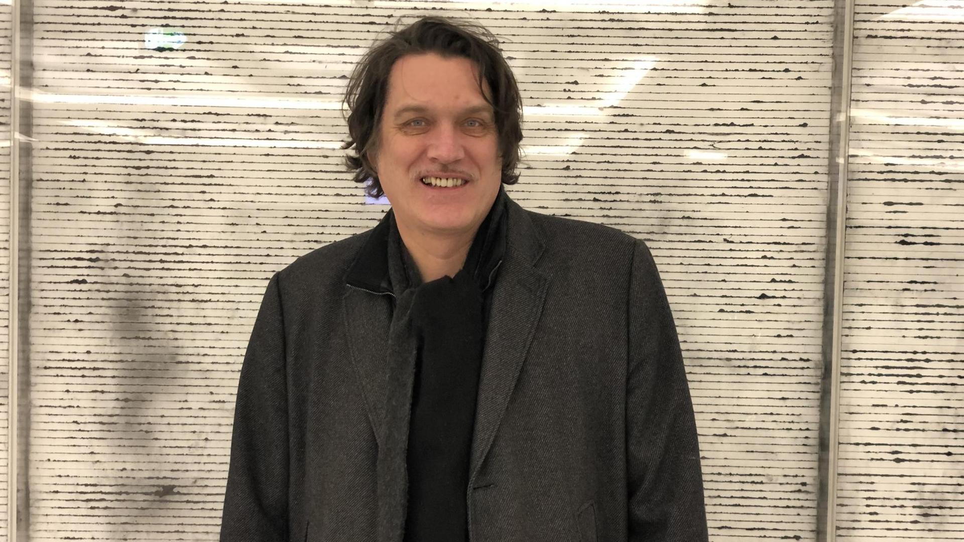 Auf dem Bild ist der Musiker Frank Spilker zu sehen, wie er vor einer grauen Wand steht und in die Kamera lächelt Auf dem Bild ist der Musiker Frank Spilker zu sehen, wie er vor einer grauen Wand steht und in die Kamera lächelt