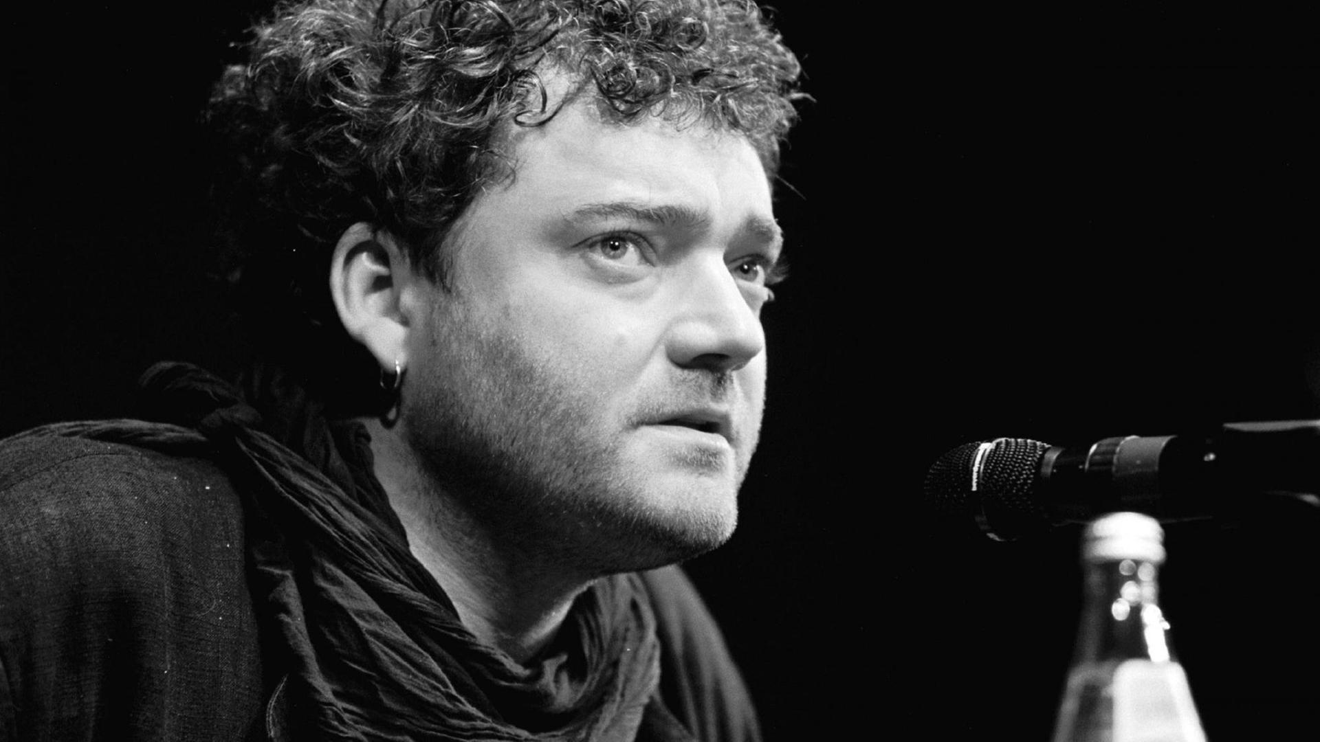 Wiglaf Droste sitzt vor einem Mikrofon auf einer Theaterbühne. Wiglaf Droste sitzt vor einem Mikrofon auf einer Theaterbühne.