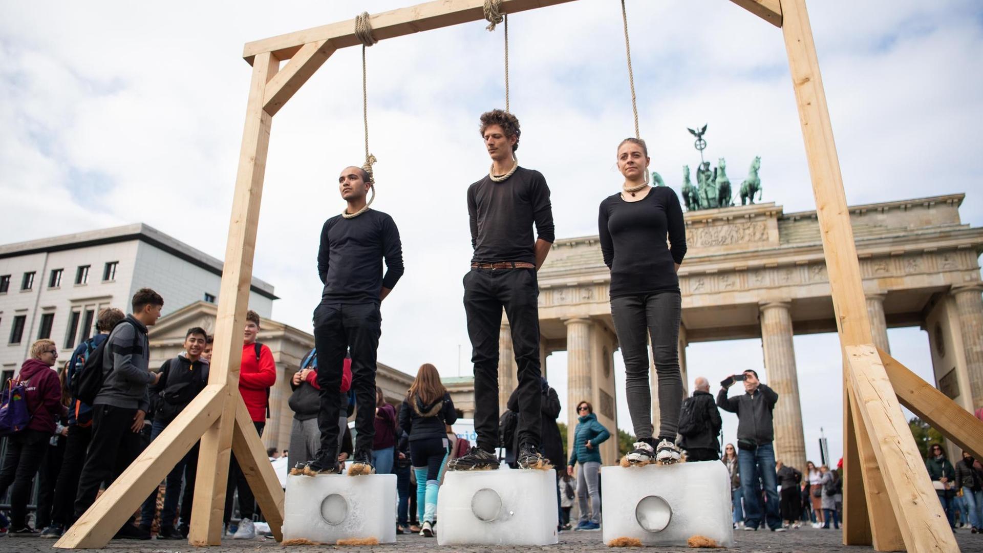 Drei Demonstranten stehen unter einem Galgen mit einem Seil um den Hals auf Eisblöcken. Dahinter ist das Brandenburger Tor. Drei Demonstranten stehen unter einem Galgen mit einem Seil um den Hals auf Eisblöcken. Dahinter ist das Brandenburger Tor.