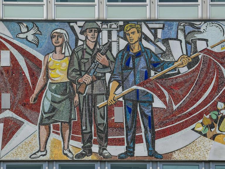Auf der Rückseite vom Haus des Lehrers in Berlin-Mitte sieht man ein Mosaikkunstwerk, das einen Mann mit Flagge, einen Soldaten und eine Frau zeigt.