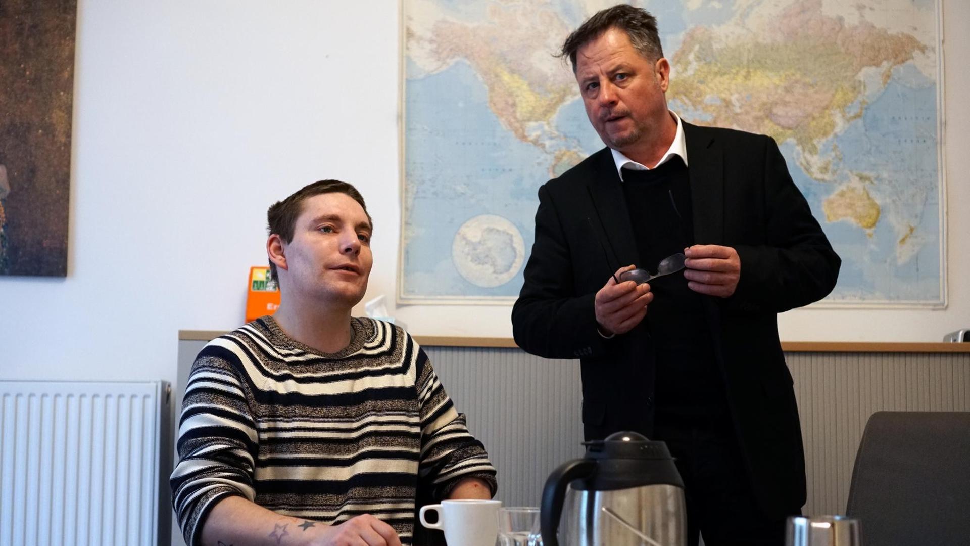 Geffert sitzt an einem Tisch, Böwe steht daneben und spricht mit ihm Geffert sitzt an einem Tisch, Böwe steht daneben und spricht mit ihm