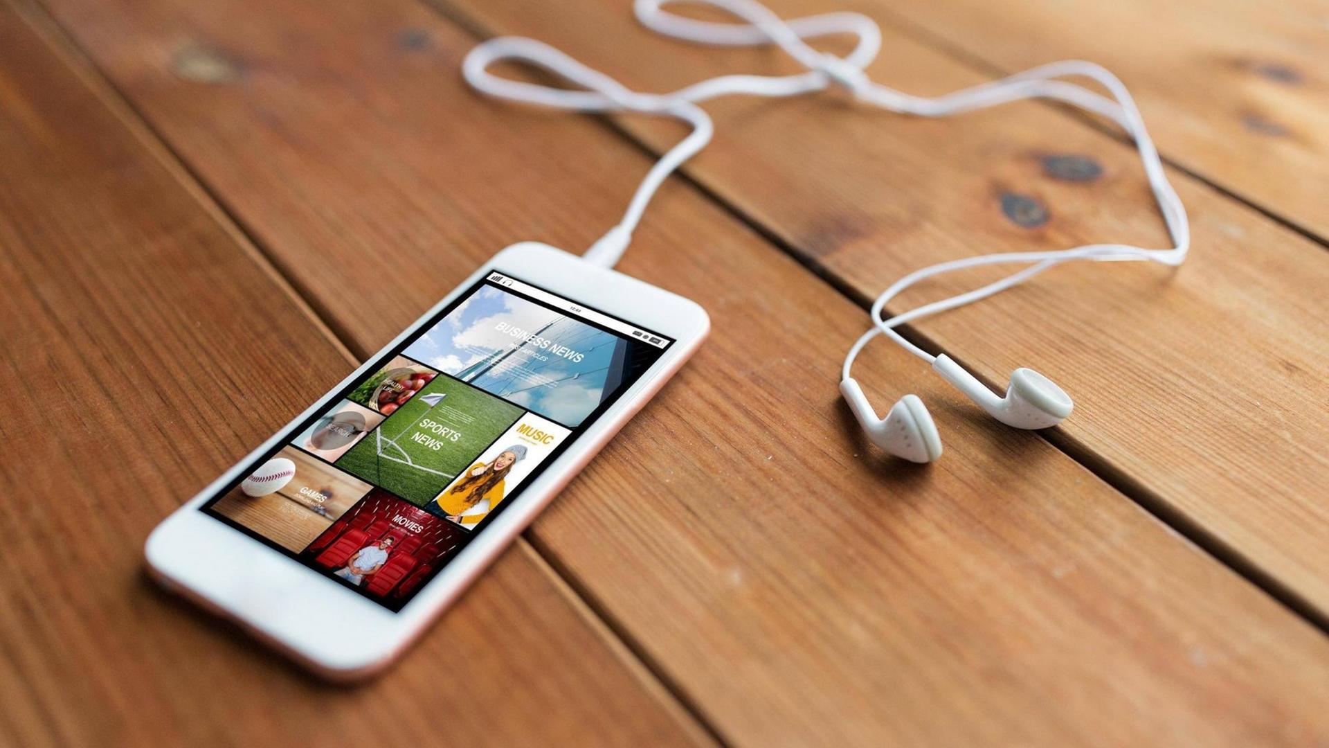 Ein Smartphone mit angeschlossenen Kopfhörern liegt auf einem Dielenboden. Auf dem Display ist die Oberfläche einer Podcastapp zu sehen. Ein Smartphone mit angeschlossenen Kopfhörern liegt auf einem Dielenboden. Auf dem Display ist die Oberfläche einer Podcastapp zu sehen.