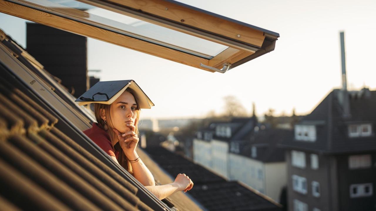 Eine junge Frau schaut nachdenklich aus einem Dachfenster mit einem aufgeschlagenen Buch über dem Kopf.