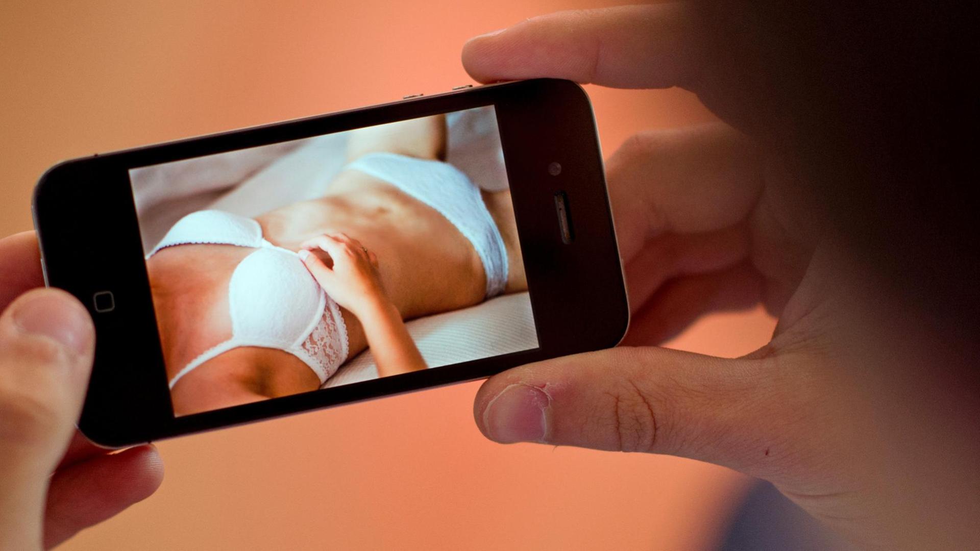 Ein Mann hält ein Smartphone in der Hand, auf dem ein erotisches Foto einer jungen Frau zu sehen ist.