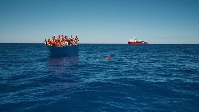 Ein überfülltes Holzboot im Mittelmeer. Im Hintergrund das deutsche Rettungsschiff "Sea Eye 4".