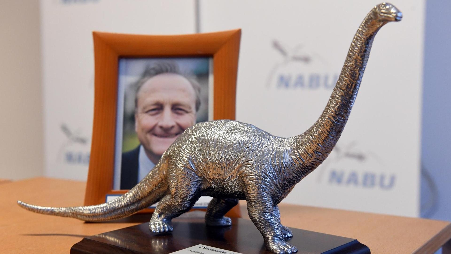 Die silbern glänzende Dinosaurier-Figur steht auf einem Tisch vor einem Foto von Bauernpräsident Rukwied