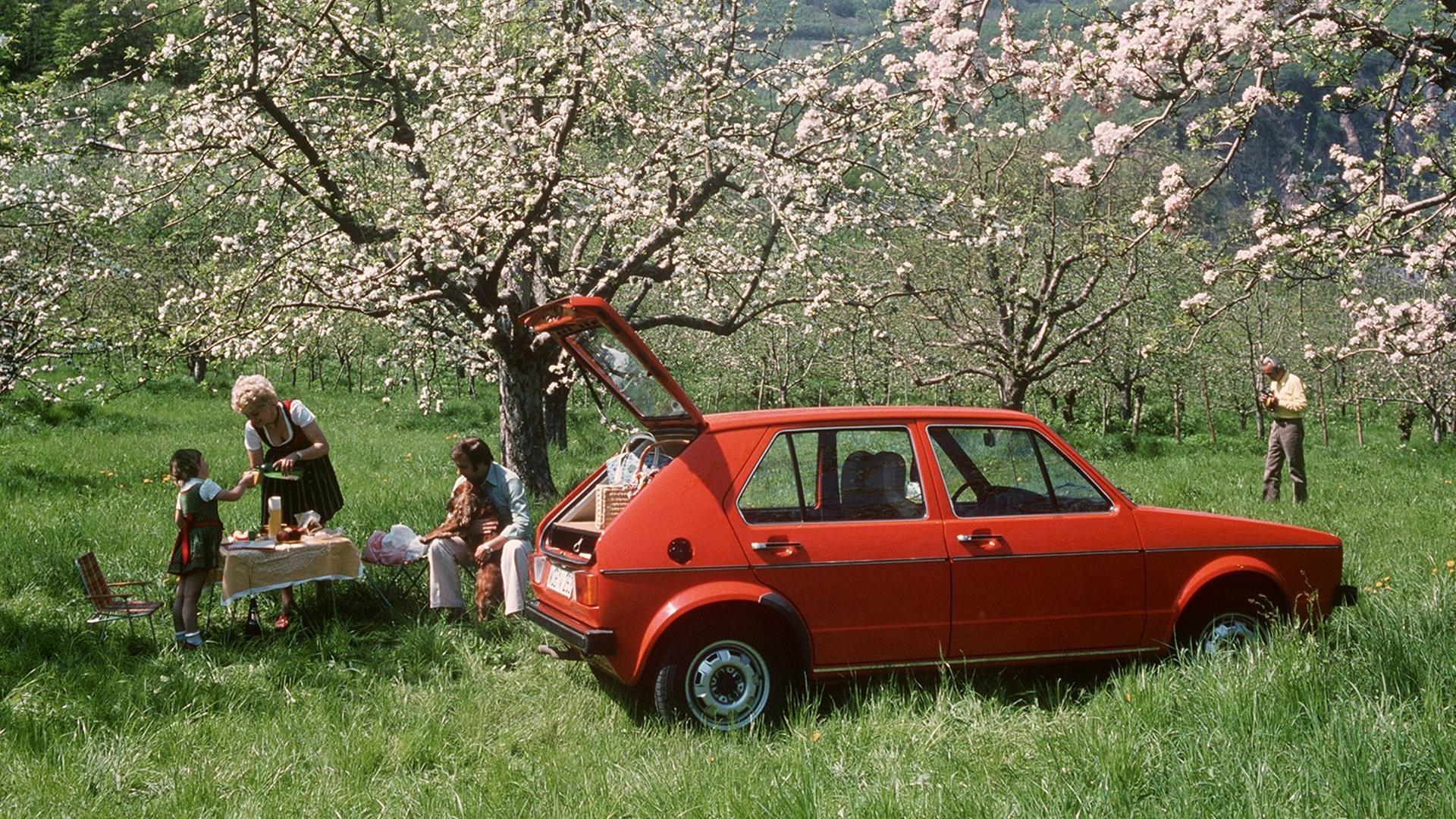 Eine Familie ist mit ihrem roten VW-Golf ins Grüne gefahren und veranstaltet ein Picknick unter blühenden Obstbäumen.