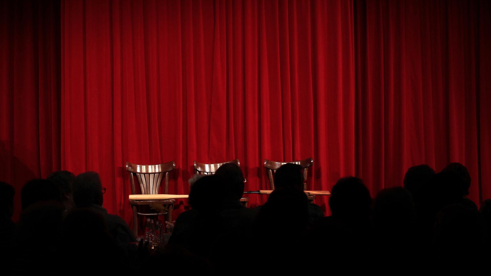 Zuschauer sitzen im Theater vor einer leeren Bühne mit geschlossenem roten Vorhang.