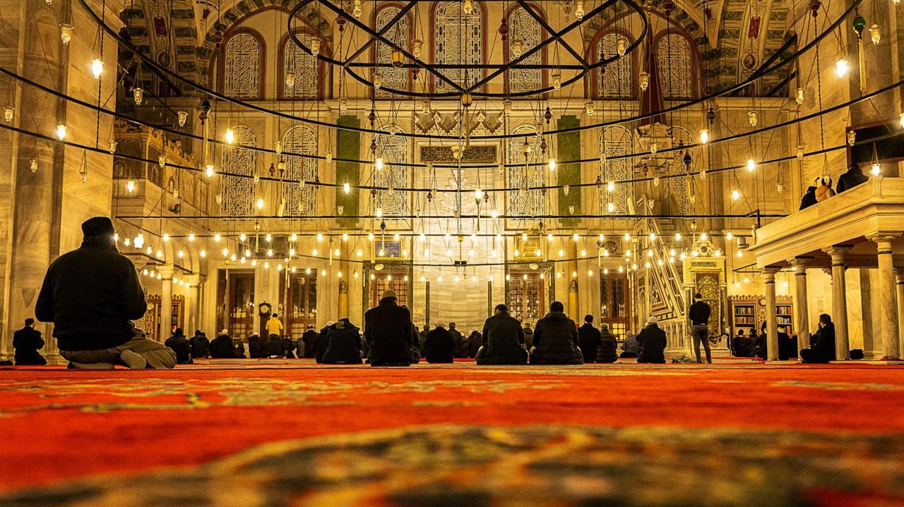 Von der Perspektive eines große roten Teppichs aus  blickt man auf die Betenden in einer großen Moschee. Schwarzgekleidete Männer, die unter vielen hängenden Glühlampen vor dem Altar sitzen.