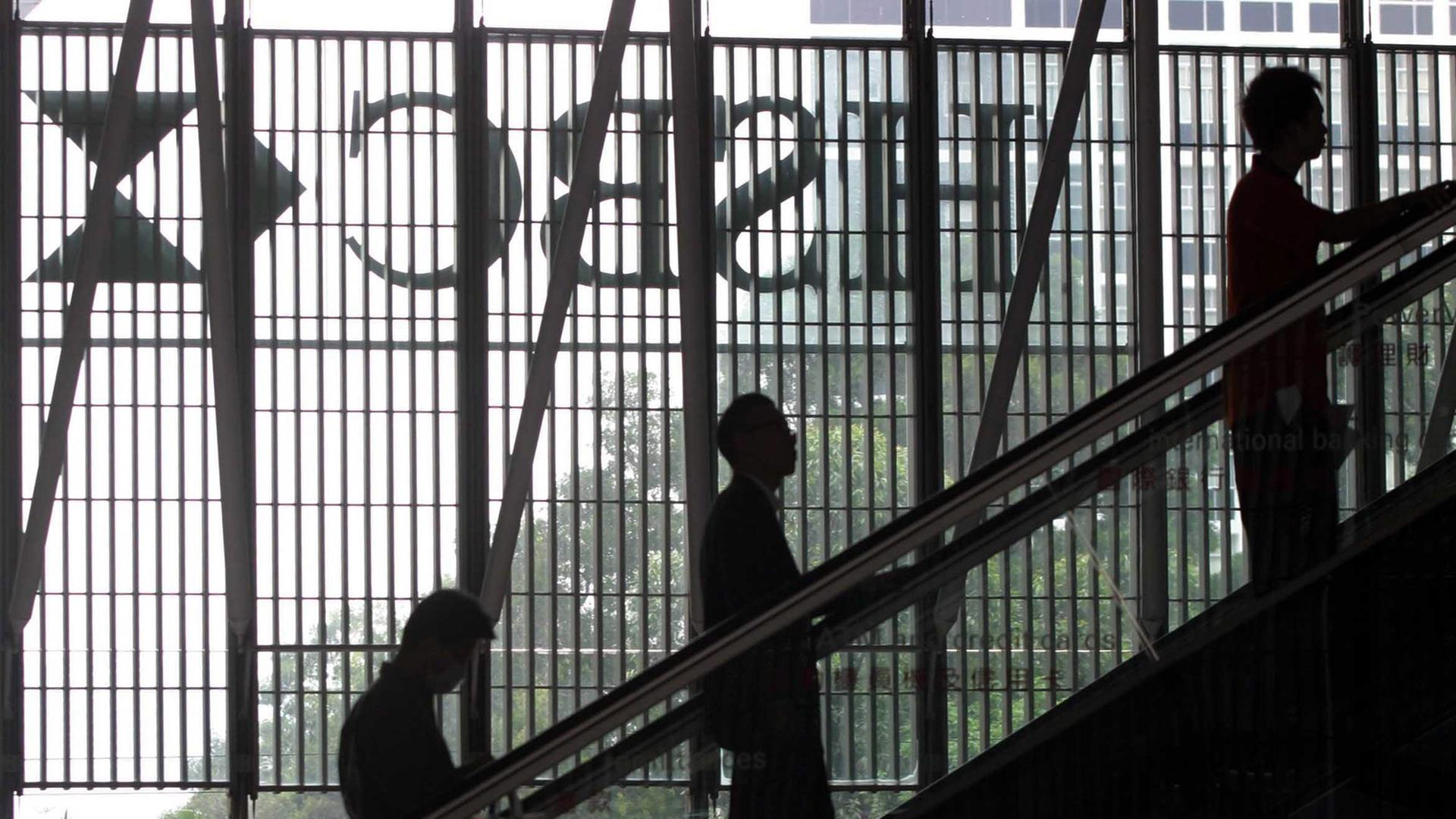 Menschen auf einer Rollstreppe, im Hintergrund das spiegelverkehrte Logo des HSBC-Bank.