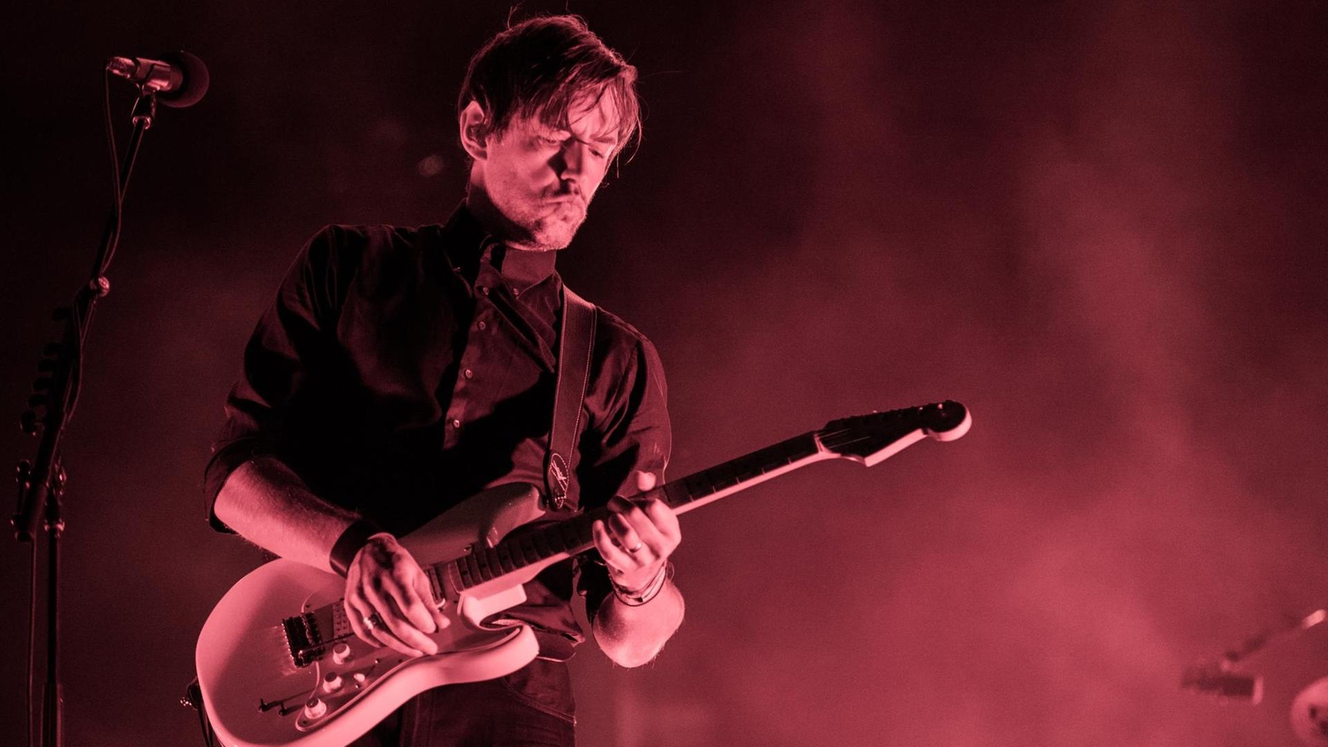 Ed O'Brien spielt E-Gitarre und wird von einem violetten Licht im Nebel beleuchtet. Ed O'Brien spielt E-Gitarre und wird von einem violetten Licht im Nebel beleuchtet.