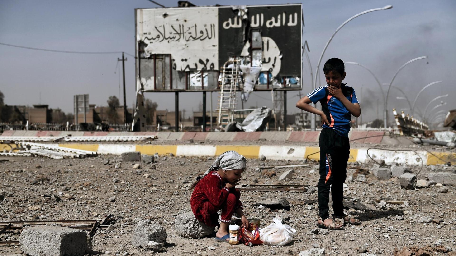 Kinder sitzen auf der Straße in Mossul - im Hintergrund eine abgerissene Fahne des IS