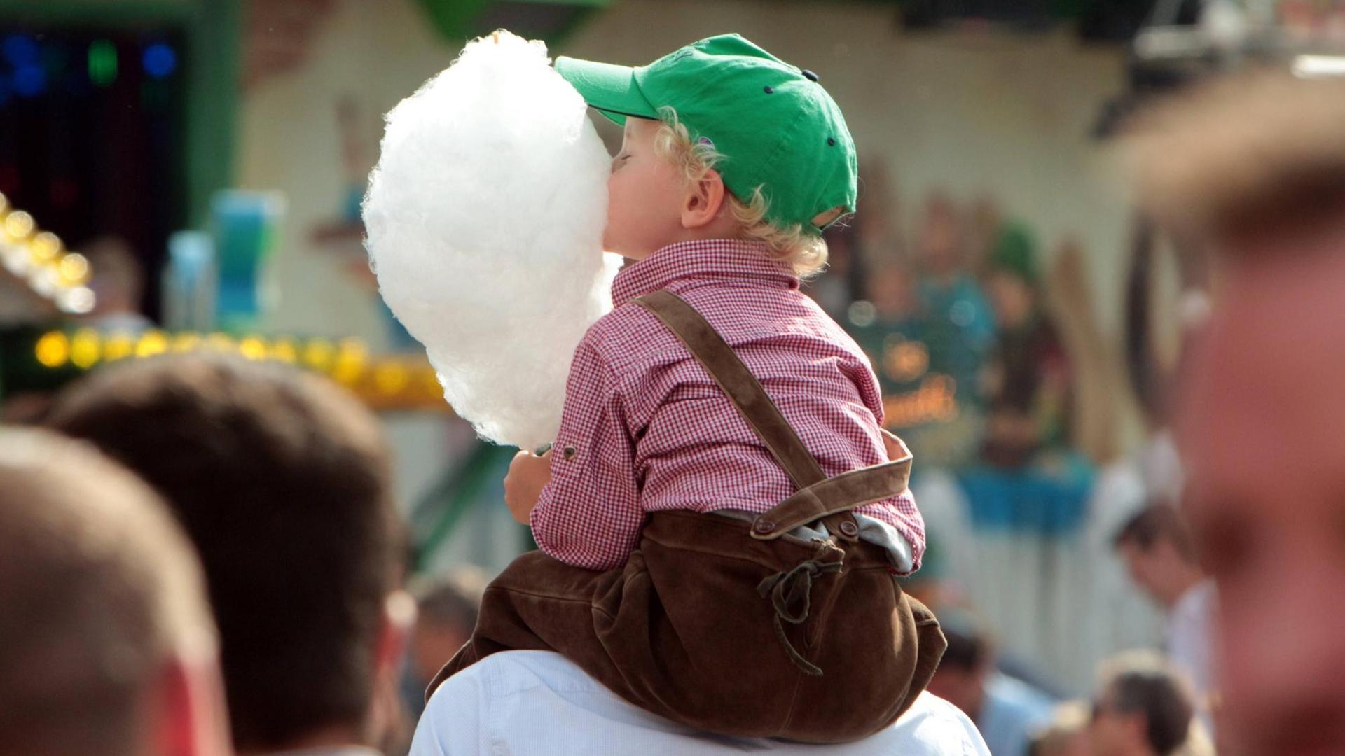 Ein Wiesn-Wochenende bei strahlendem Sonnenschein: Vater in Tracht trägt seinen kleinen Sohn, der beherzt in seine Zuckerwatte beißt, auf den Schultern. Ein Wiesn-Wochenende bei strahlendem Sonnenschein: Vater in Tracht trägt seinen kleinen Sohn, der beherzt in seine Zuckerwatte beißt, auf den Schultern.