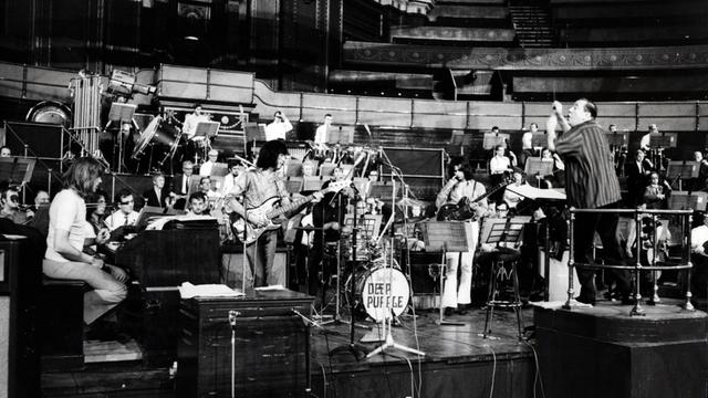Schwarz-weiß Foto von der Band Deep Purple und Mitgliedern des Royal Philharmonic Orchestra in der Royal Albert Hall. Mitglieder der Rockband und Sir Malcolm Arnold tragen legere Kleidung, die Mitglieder des Orchester Anzug mit Schlips, mit und ohne Jacket