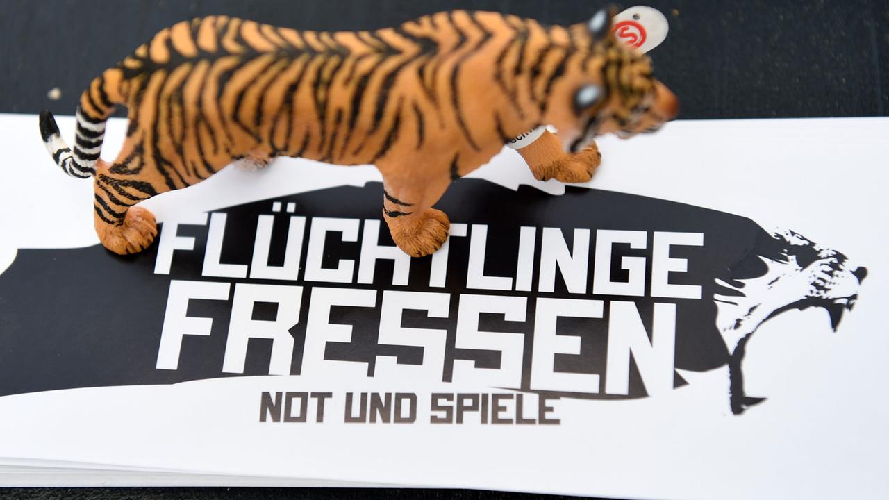 Ein schwarz-weißer Aufkleber mit der Aufschrift "Flüchtlinge fressen - Not und Spiele", auf dem eine Tigerfigur steht. Ein schwarz-weißer Aufkleber mit der Aufschrift "Flüchtlinge fressen - Not und Spiele", auf dem eine Tigerfigur steht.