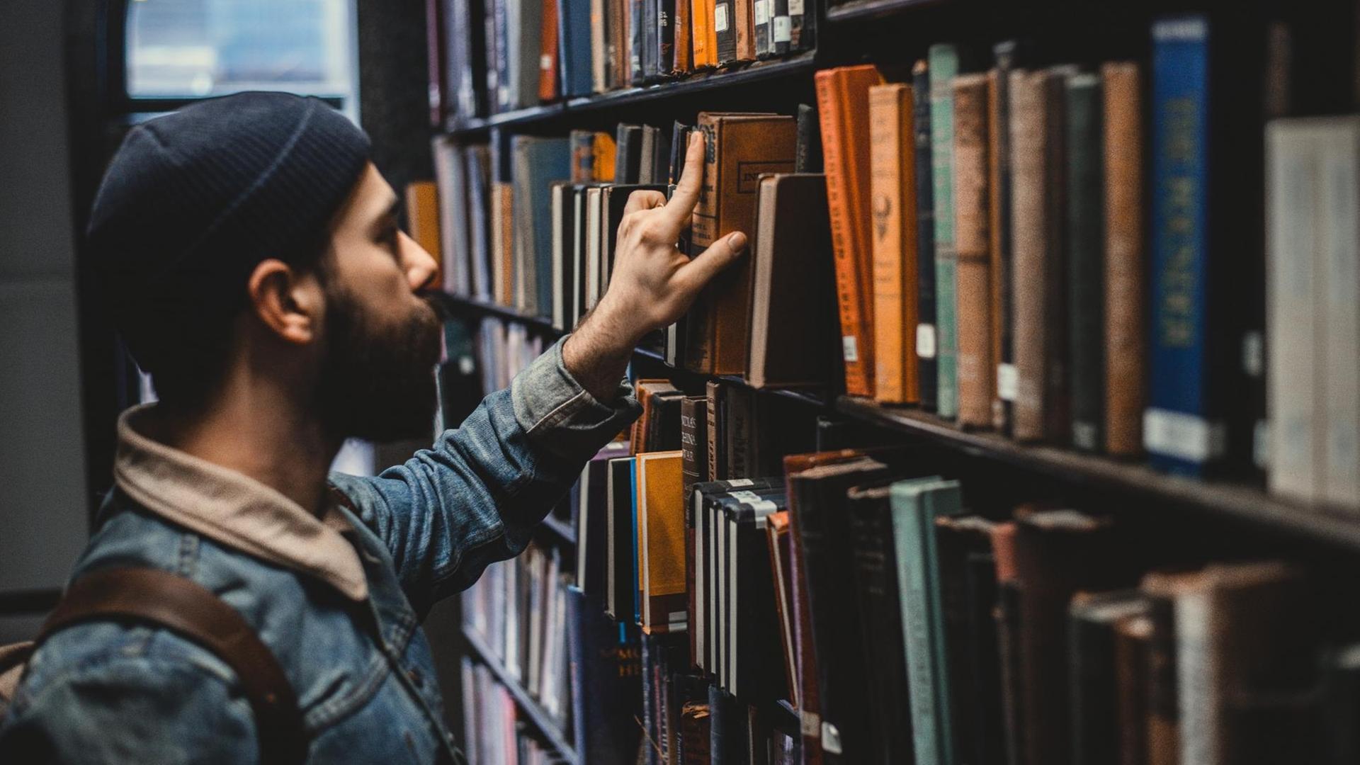 Ein junger Mann zieht ein gebundenes altes Buch aus einem Bibliotheksregal. Ein junger Mann zieht ein gebundenes altes Buch aus einem Bibliotheksregal.