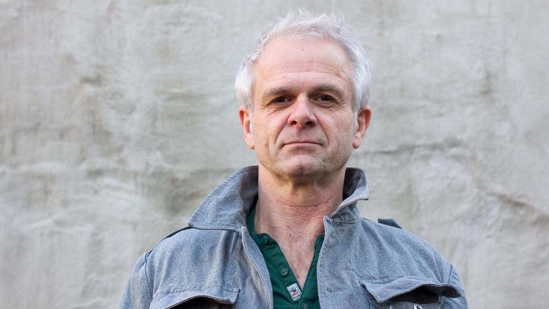 Ein Mann mit weißem Haar schaut auf einer Porträtaufnahme direkt in die Kamera, er steht vor einer grauen Wand und trägt eine Jacke aus hellblauem Stoff, darunter ein grünes Hemd. Ein Mann mit weißem Haar schaut auf einer Porträtaufnahme direkt in die Kamera, er steht vor einer grauen Wand und trägt eine Jacke aus hellblauem Stoff, darunter ein grünes Hemd.