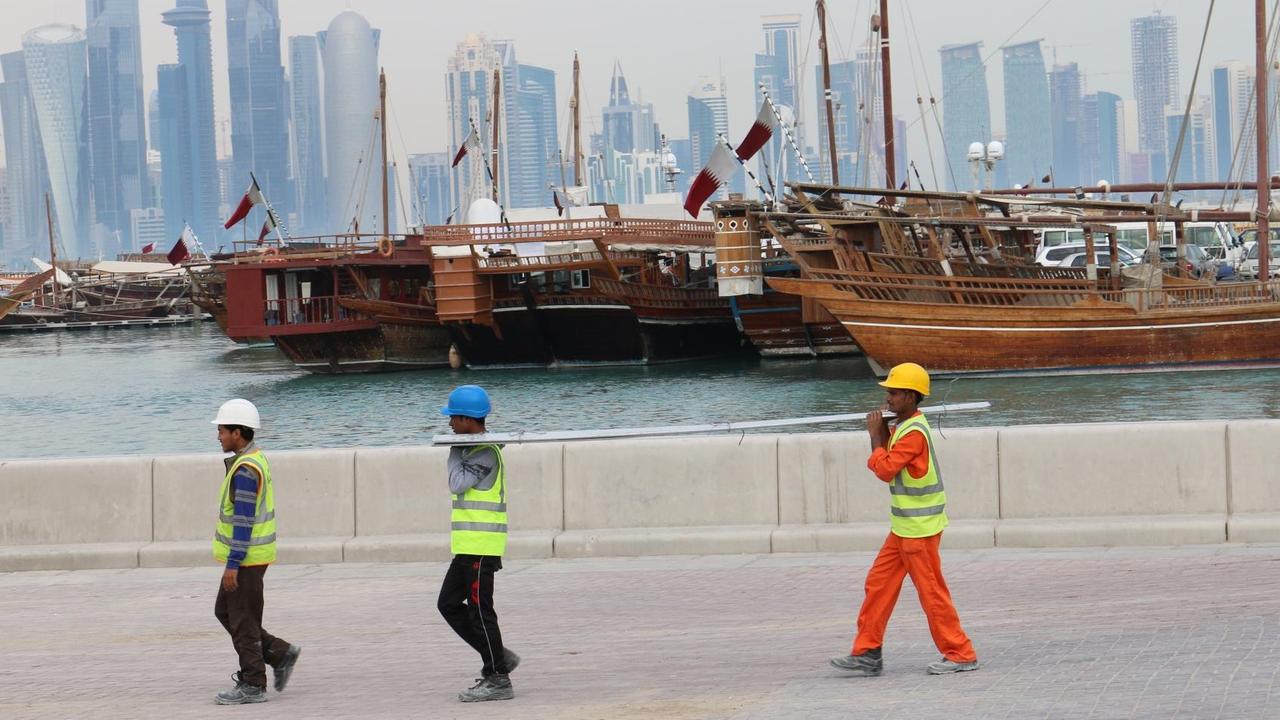 Drei Bauarbeiter gehen am Dau-Hafen entlang mit der Skyline der katarischen Hauptstadt Doha im Hintergrund. Drei Bauarbeiter gehen am Dau-Hafen entlang mit der Skyline der katarischen Hauptstadt Doha im Hintergrund.