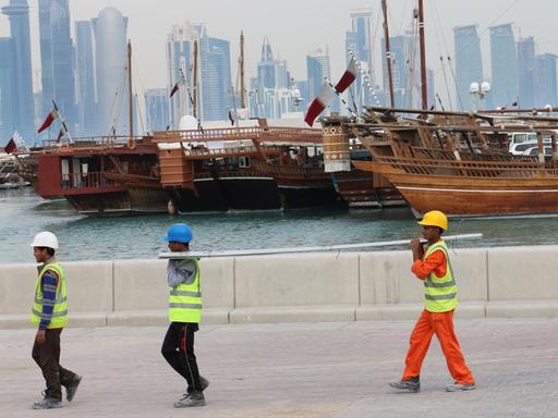 Drei Bauarbeiter gehen am Dau-Hafen entlang mit der Skyline der katarischen Hauptstadt Doha im Hintergrund.