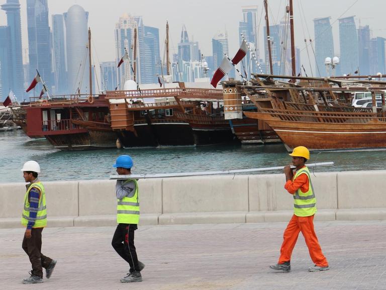 Drei Bauarbeiter gehen am Dau-Hafen entlang mit der Skyline der katarischen Hauptstadt Doha im Hintergrund.