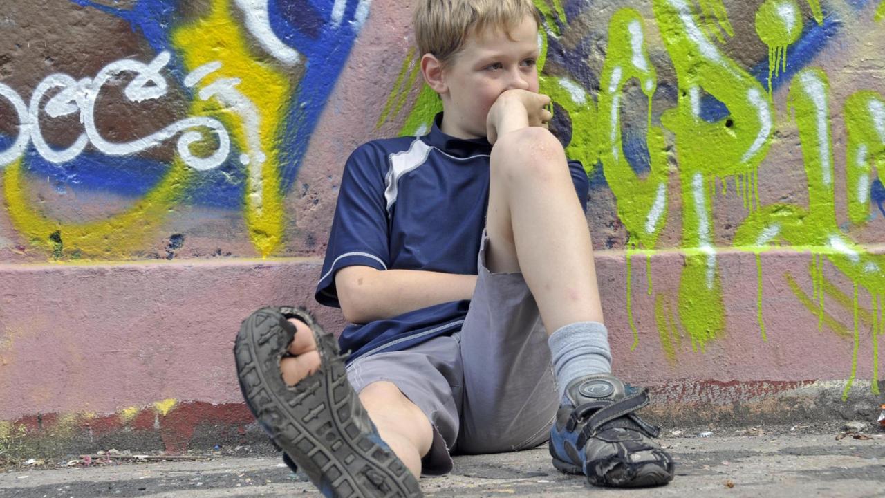 Lustloser neunjähriger Junge sitzt vor einer Graffiti-Wand, einen Fuß von sich gestreckt, sodass die kaputte Sohle des Schuhs sichtbar wird.