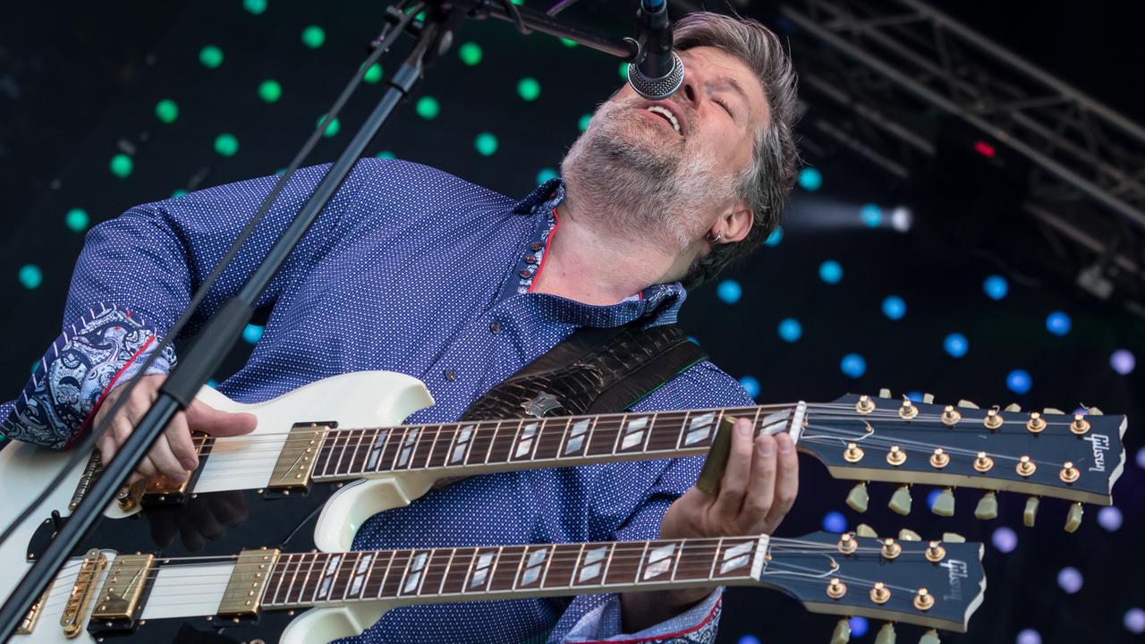 Ein Mann im blauen Hemd steht auf einer Bühne und spielt eine doppelhalsige Gitarre.