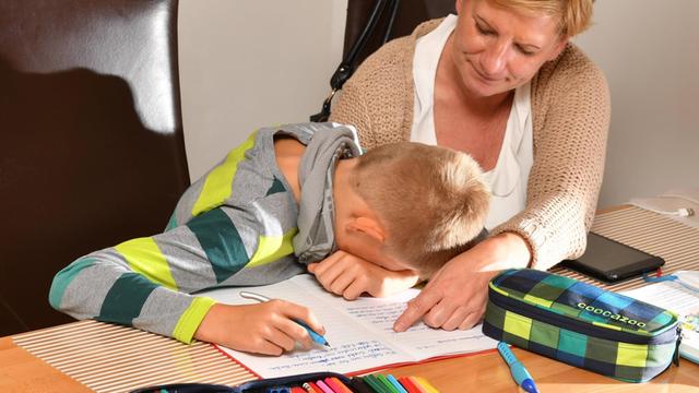 Eine Mutter hilft ihrem Sohn bei den Schulaufgaben, der verzweifelt den Kopf auf sein Heft gelegt hat.