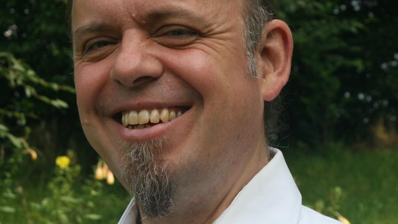 Porträt des Pfarrers und Übersetzers Matthias Binder, lächelnd, mit offenem weißen Hemd, im Garten Porträt des Pfarrers und Übersetzers Matthias Binder, lächelnd, mit offenem weißen Hemd, im Garten