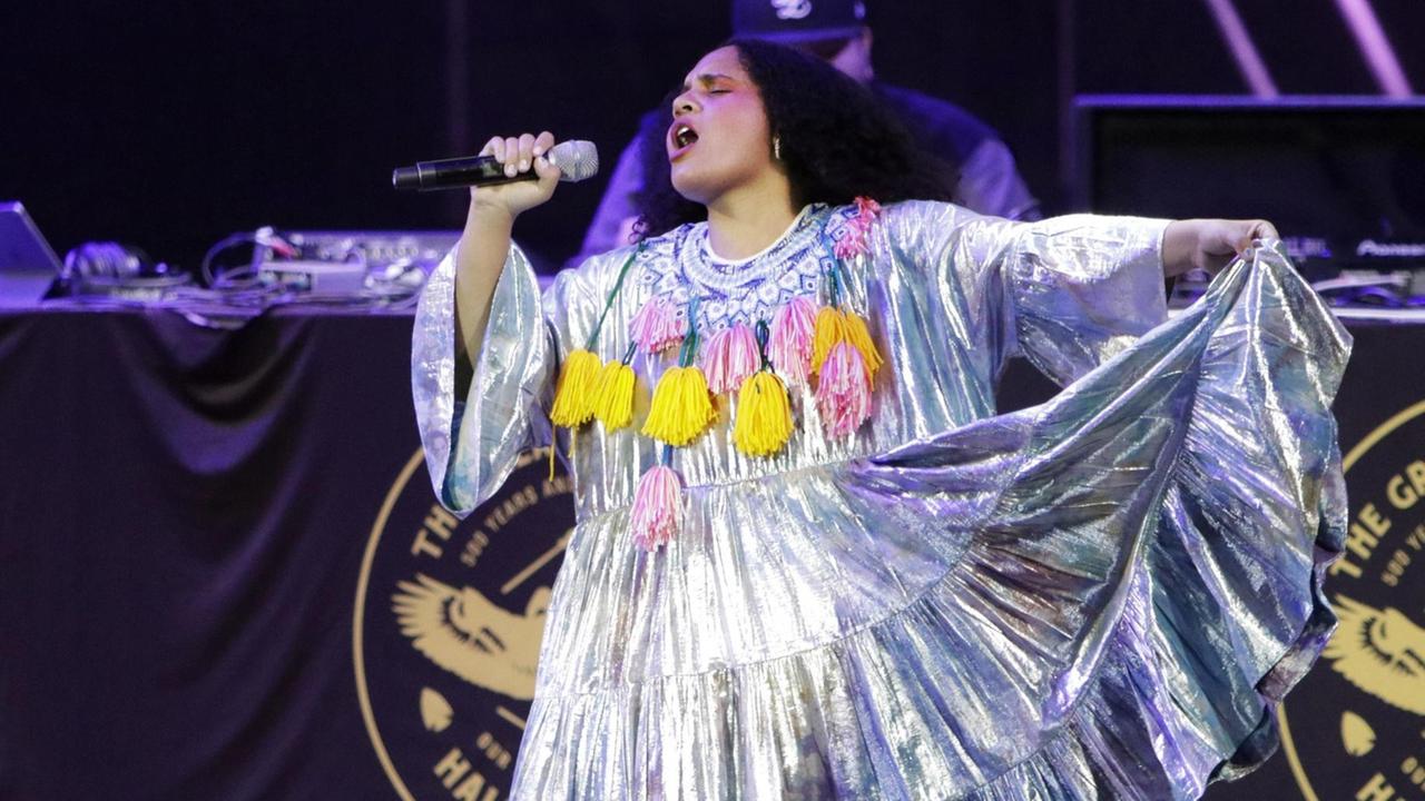 Lido Pimienta singend auf der Bühne, in einem langen, silber schimmernden Kleid. Lido Pimienta singend auf der Bühne, in einem langen, silber schimmernden Kleid.