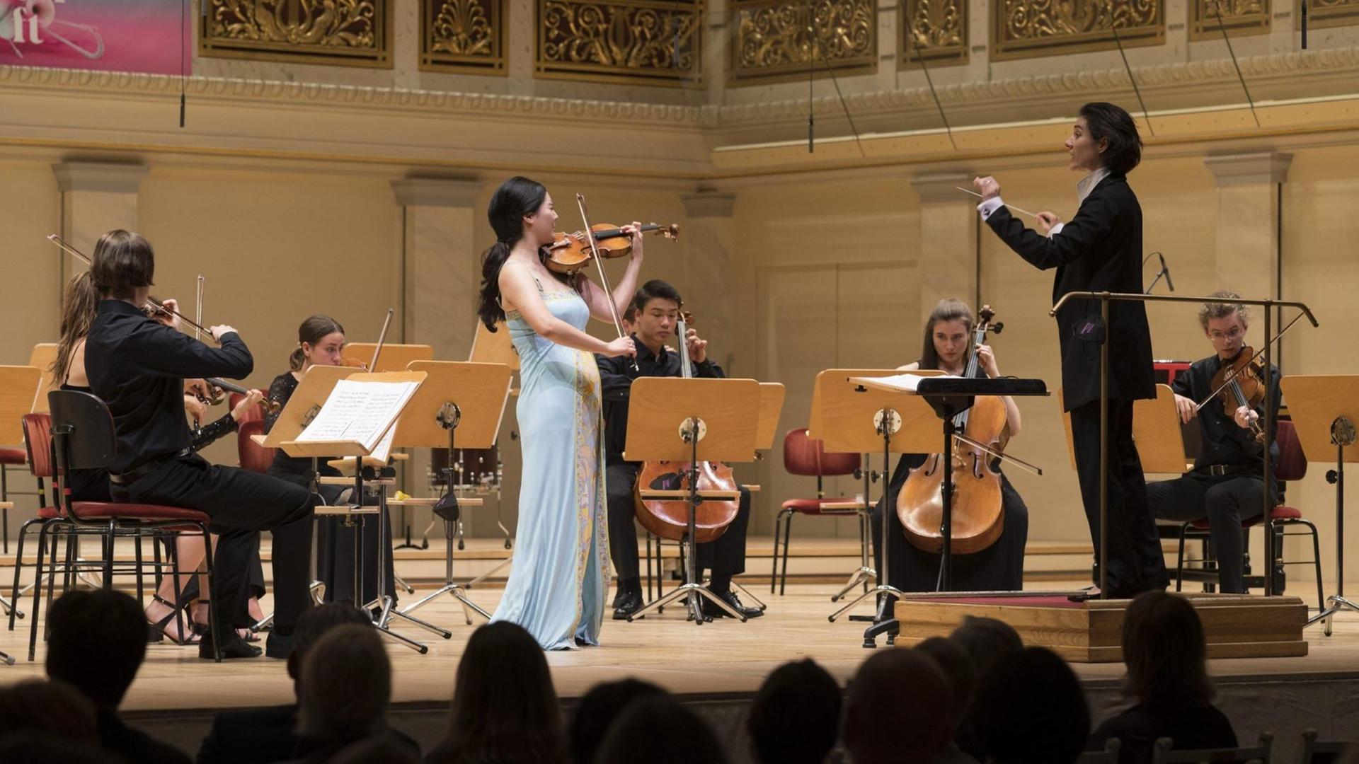 Eine junge Geigerin steht als Solistin vor einem Jugendorchester auf der Bühne im Konzerthaus Berlin. Am Pult eine junge Dirigentin im schwarzen Anzug. Eine junge Geigerin steht als Solistin vor einem Jugendorchester auf der Bühne im Konzerthaus Berlin. Am Pult eine junge Dirigentin im schwarzen Anzug.
