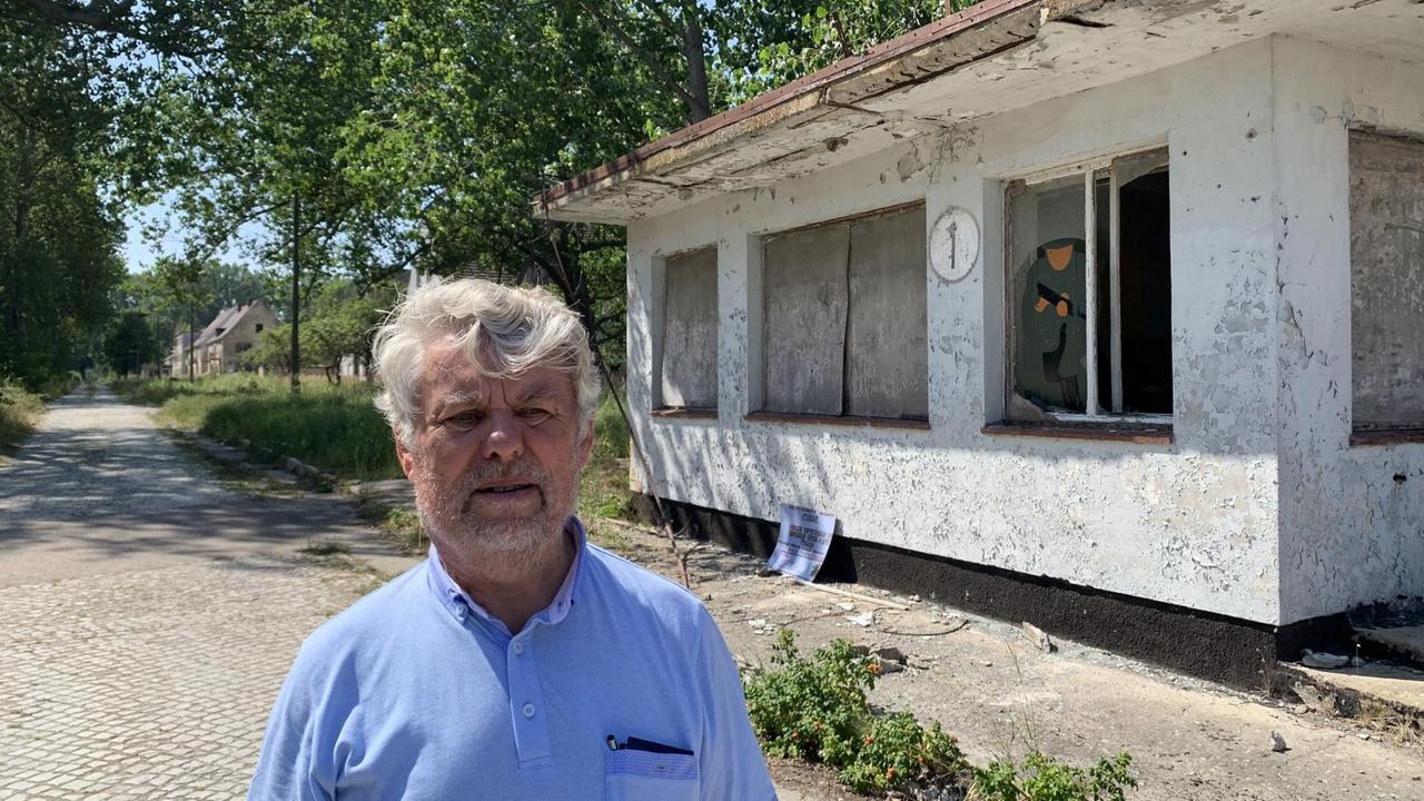 Klaus Feiler steht vor der Ruine eines weißen Flachbaus. Hinter ihm verläuft eine Kopfsteinpflasterstraße. Klaus Feiler steht vor der Ruine eines weißen Flachbaus. Hinter ihm verläuft eine Kopfsteinpflasterstraße.