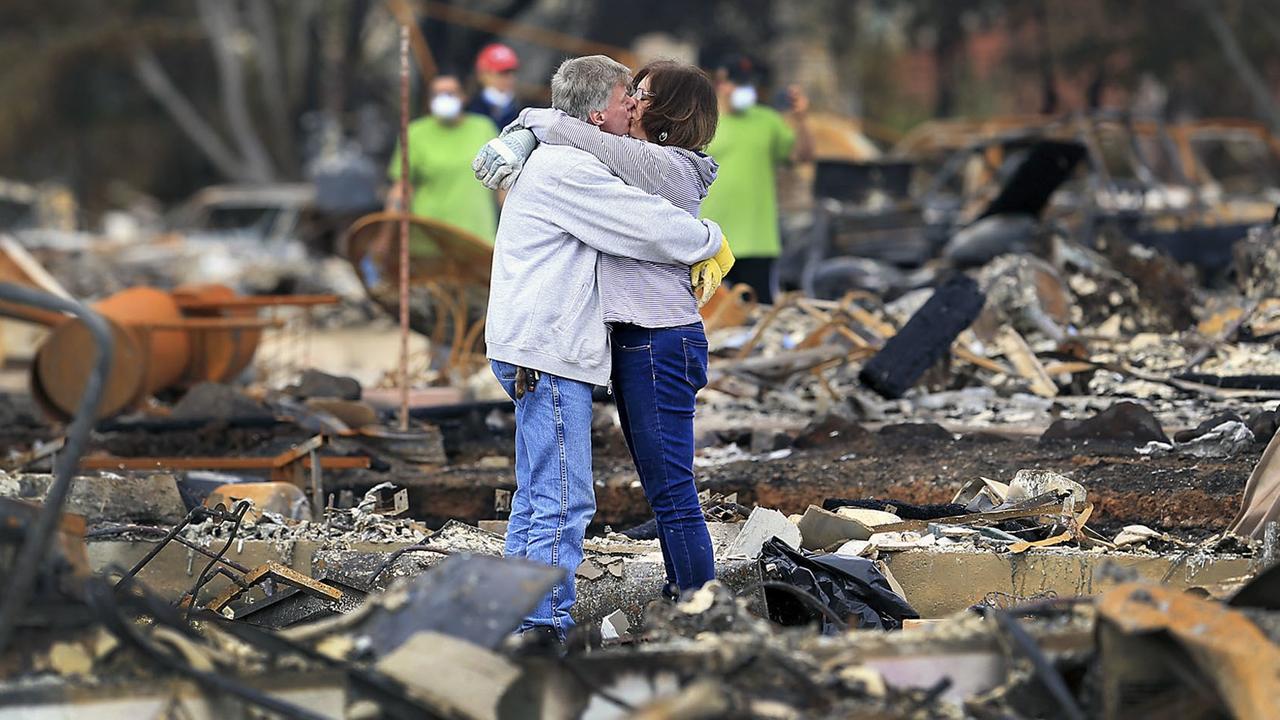 Die beiden stehen engumschlungen inmitten der verkohlten Überreste. Im Hintergrund unscharf weitere Menschen mit Atemschutzmasken. Die beiden stehen engumschlungen inmitten der verkohlten Überreste. Im Hintergrund unscharf weitere Menschen mit Atemschutzmasken.