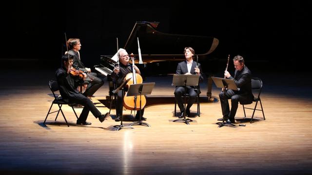 Fünf Musiker sitzen mit ihren Instrumenten auf einer ins Scheinwerferlicht getauchten Bühne.