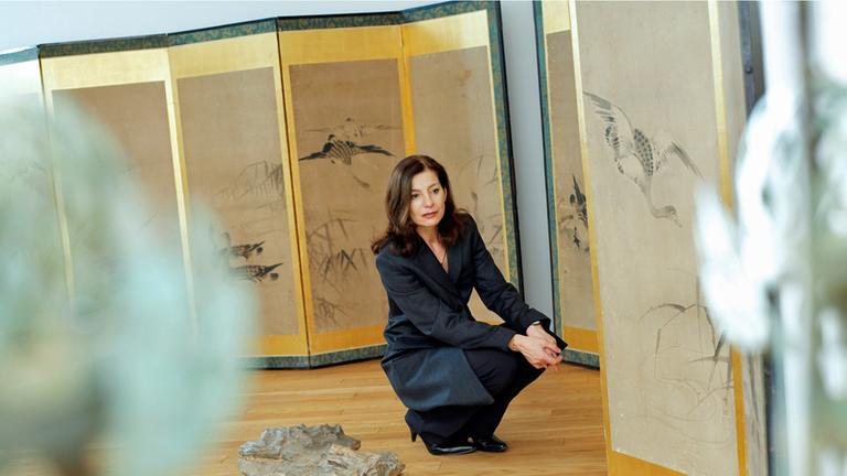 Die Schriftstellerin Ursula Krechel hockt gedankenversunken in einer Ausstellung. Auf Stellwänden sind Zeichnungen von Vögeln und Gräsern zu sehen.