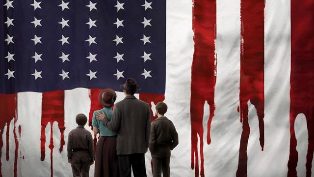 Eine Familie von hinten vor einer US-amerikansichen Flagge. Die Roten STreifen laufen herab, wie Blut.