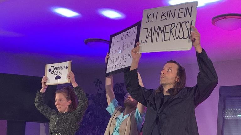 Rika Weniger, Noah Voelker und Burkhard Körner halten auf der Bühne Schilder hoch mit der Aufschrift "Ich bin ein Jammerossi".