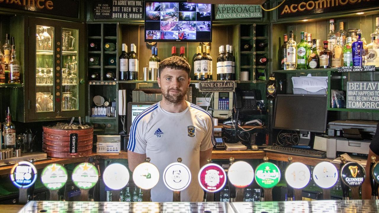 Tom Quinn im Traditionspub "Ferryman" in den Docks von Dublin. Er hat kurze Haare, ein Fußball-Trikot an und steht vor den Zapfhähnen. Tom Quinn im Traditionspub "Ferryman" in den Docks von Dublin. Er hat kurze Haare, ein Fußball-Trikot an und steht vor den Zapfhähnen.