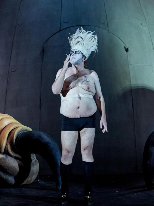 Ein Mann in Unterhose und einem Federkopfschmuck steht mit weiß geschminktem Gesicht auf einer Theaterbühne, raucht und sieht dabei nachdenklich in die Ferne. Neben ihm zwei weitere leicht gekleidete Personen, sie stehen zwischen riesigen Krallenfüßen.