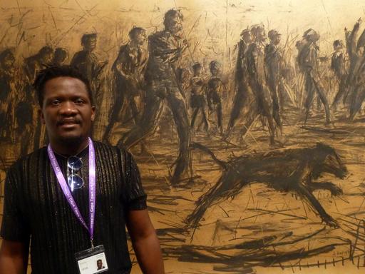 Der Maler Asanda Kupa vor einem seiner Werke, einer Zeichnung einer nach rechts gehenden Menschenmenge mit einem Hund im Vordergrund, die wahrscheinlich mit Kohle gezeichnet wure, bei der Kunstmesse in Kapstadt