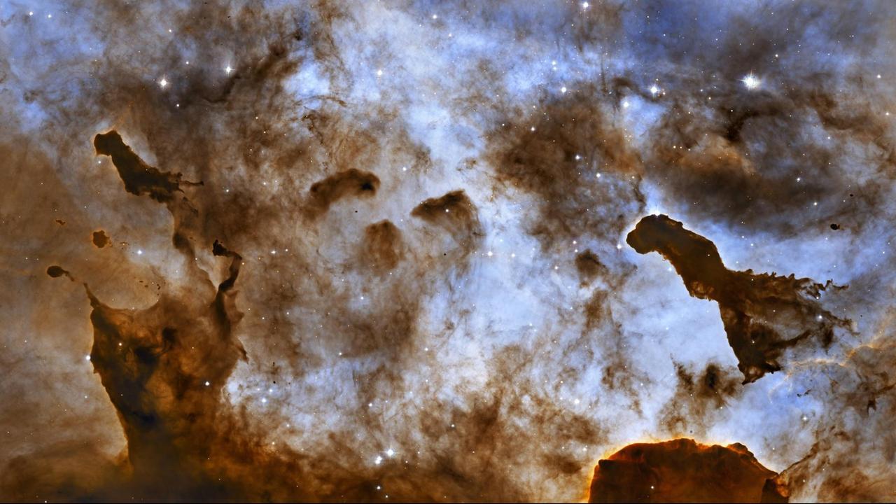 Eine Aufnahme der Pillars of Creation: Zu sehen sind undefinierbare braune Strukturen in einem hellen Nebel, es leuchten Sterne auf hellblauen Hintergrund.