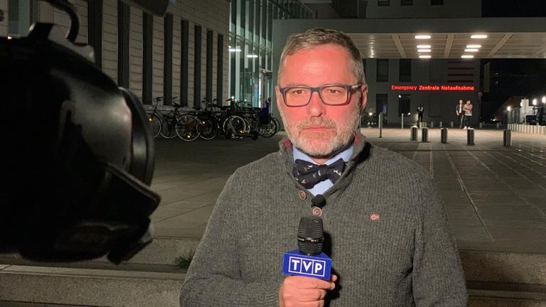 Ein Mann steht mit Wolljacke, Fliege und einem TVP-Mikrofon in der Hand vor einer Kamera. Hinter ihm die Notaufnahme eines Krankenhauses. Ein Mann steht mit Wolljacke, Fliege und einem TVP-Mikrofon in der Hand vor einer Kamera. Hinter ihm die Notaufnahme eines Krankenhauses.
