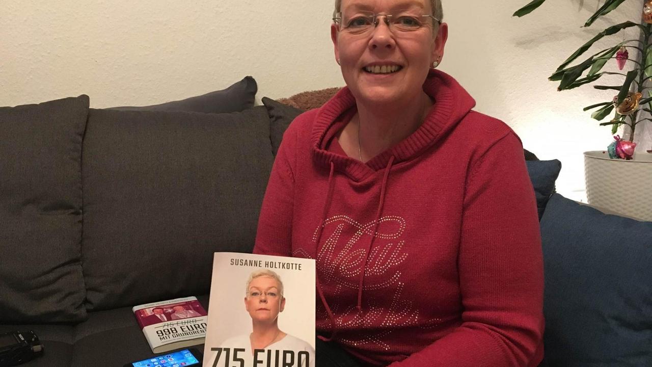 Susanne Holtkotte sitzt auf einer Couch und hält ein Buch mit dem Titel "715 Euro" in der Hand