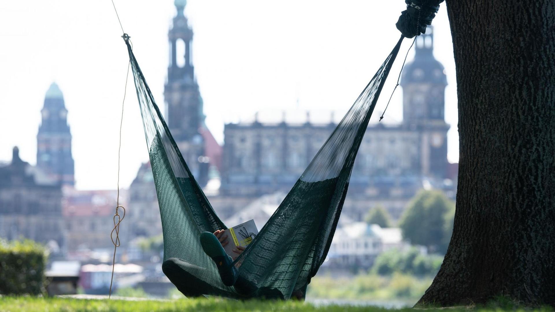 Ein Mann liegt in einer Hängematte und liest ein Buch vor der Kulisse der Altstadt mit dem Rathausturm, der Katholischen Hofkirche und dem Hausmannsturm. Ein Mann liegt in einer Hängematte und liest ein Buch vor der Kulisse der Altstadt mit dem Rathausturm, der Katholischen Hofkirche und dem Hausmannsturm.