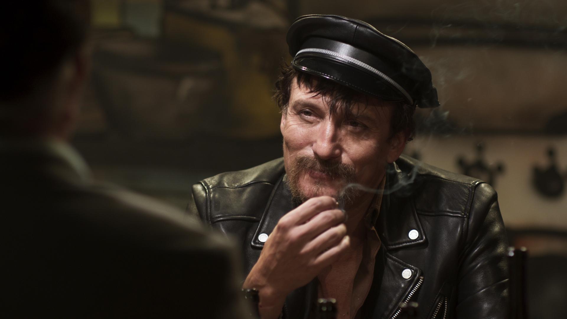 Szene aus dem Film "Enfant Terrible": Rainer Werner Fassbinder (gespielt von Oliver Masucci) in schwarzer Lederjacke und mit Polizeimütze. Szene aus dem Film "Enfant Terrible": Rainer Werner Fassbinder (gespielt von Oliver Masucci) in schwarzer Lederjacke und mit Polizeimütze.